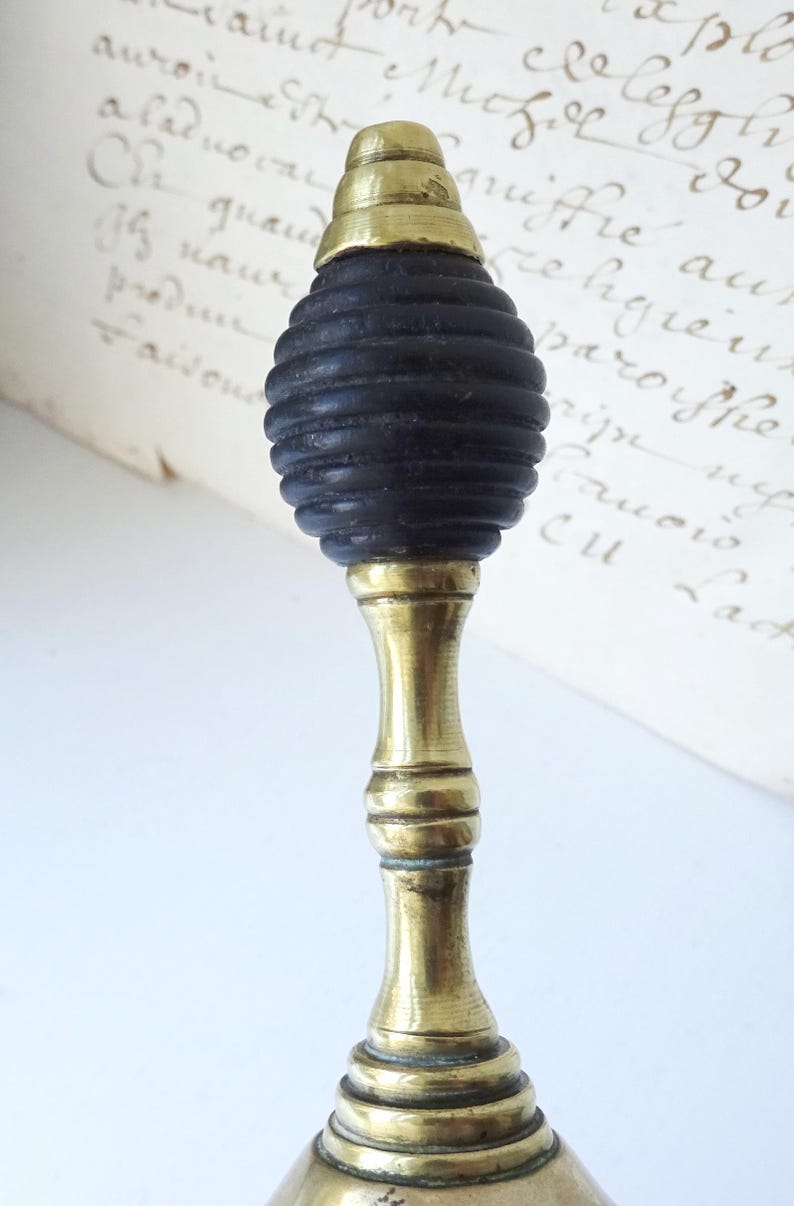 Puede incluir: Un tap&oacute;n de botella vintage con un v&aacute;stago de lat&oacute;n y una parte superior negra y acanalada. La parte superior est&aacute; rematada con un acento de lat&oacute;n. El tap&oacute;n se encuentra sobre un fondo de papel envejecido con escritura manuscrita.