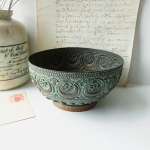 May include: An antique bronze bowl with intricate floral and crescent moon patterns. The bowl has a weathered, verdigris finish and sits on a small wooden base. The bowl is a decorative item.