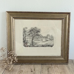 May include: A framed black and white landscape print. The artwork depicts a detailed scene with trees, a building, and figures. The frame is a gold color, and the print is displayed on a wooden surface.