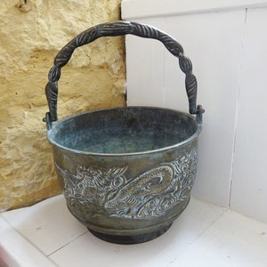 Può includere: Secchio antico in bronzo con un rilievo decorativo a forma di drago. Il secchio ha un manico intrecciato e una base rotonda. Il design del drago è dettagliato e aggiunge un tocco unico al pezzo. Il secchio è probabilmente in ottone o metallo simile.