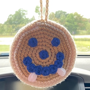 May include: A crocheted smiley face hanging from a rearview mirror. The face is brown with blue eyes and a blue smile. The smile is outlined with pink.