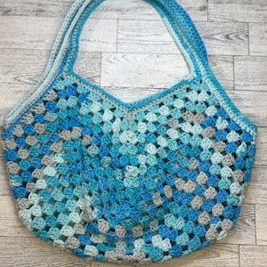 May include: A handmade crochet tote bag featuring a classic granny square pattern in various shades of blue, light blue, and grey. The bag has a wide, open top and two integrated shoulder straps, all constructed with a consistent yarn texture.