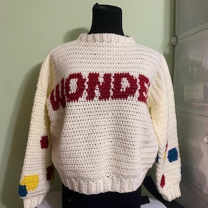 May include: Cream-colored, crocheted sweater with the word "WONDER" in red across the chest. The sleeves have colorful polka dots in red, yellow, and blue. The sweater is displayed on a black mannequin.