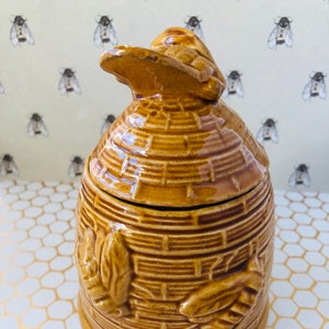 May include: A ceramic honey pot in the shape of a beehive. The pot is a warm brown colour with a textured, woven design. The lid has a small, curved handle. Decorative leaves and bees are embossed on the sides.