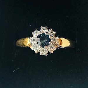 May include: A gold ring with a dark blue center stone surrounded by smaller white stones. The ring is set in a black box with a white patterned exterior.