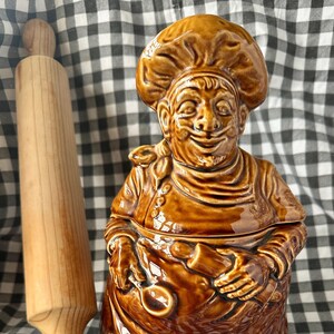 May include: A brown ceramic flour jar in the shape of a chef, with the word "FLOUR" on the front. A wooden rolling pin is next to the jar. The jar has a lid and is on a checkered background.
