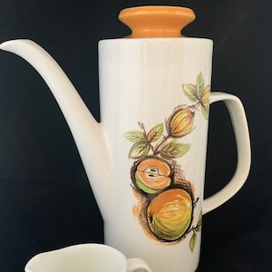 Puede incluir: Un juego de cafetera y crema de cerámica blanca. La cafetera tiene un pico largo y una tapa naranja. Ambas piezas están decoradas con un diseño de frutas en tonos naranja, verde y marrón. La crema tiene un asa pequeña.