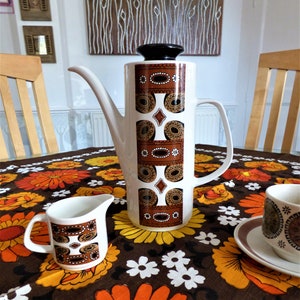 May include: A white ceramic coffee pot with a black lid and a brown and cream geometric pattern. A matching creamer and cup and saucer sit on a floral tablecloth. The table is set for coffee.