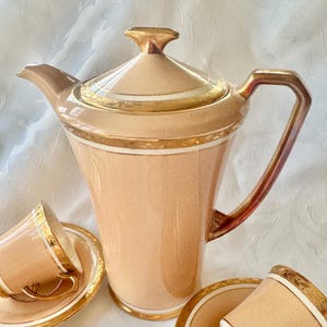K&ouml;nnte beinhalten: Pfirsichfarbenes Teeservice mit goldfarbenen Verzierungen. Das Set umfasst eine hohe, elegante Teekanne mit Deckel und Griff sowie mehrere Teetassen mit Untertassen. Die goldfarbenen Verzierungen betonen die R&auml;nder der Teekanne, Tassen und Untertassen.