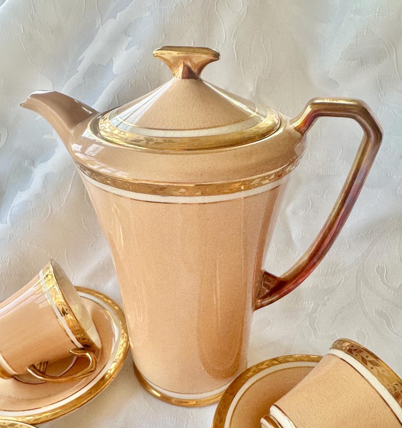 K&ouml;nnte beinhalten: Pfirsichfarbenes Teeservice mit goldfarbenen Verzierungen. Das Set umfasst eine hohe, elegante Teekanne mit Deckel und Griff sowie mehrere Teetassen mit Untertassen. Die goldfarbenen Verzierungen betonen die R&auml;nder der Teekanne, Tassen und Untertassen.