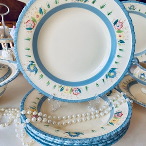 May include: A stack of cream-colored ceramic plates with a light blue border and floral design. The plates have a scalloped edge and are decorated with blue, yellow, and pink flowers. A pearl necklace is draped across the plates.