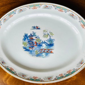 May include: Oval serving platter with a detailed Asian-inspired landscape scene in the center. The white ceramic platter features a blue, red, and green pagoda, bridge, and trees. The rim has a decorative border with gold trim.
