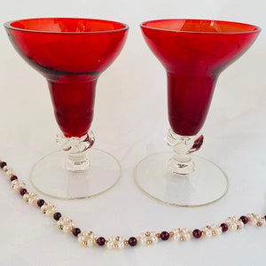 May include: Two ruby red wine glasses with clear bases and a decorative twist near the bowl. A beaded necklace with white pearls, gold beads, and dark red beads is in the foreground.