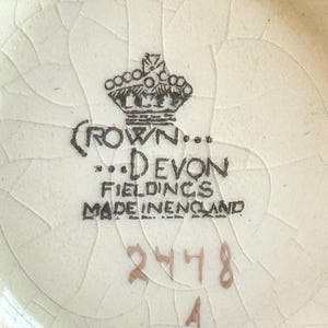 K&ouml;nnte beinhalten: Nahaufnahme einer cremefarbenen Keramikoberfl&auml;che mit Craquel&eacute;-Glasur. Das Bild zeigt den Text "CROWN... DEVON FIELDINGS MADE IN ENGLAND", die Zahlen "2478" und den Buchstaben "A". Ein schwarzes Kronenlogo ist ebenfalls zu sehen.