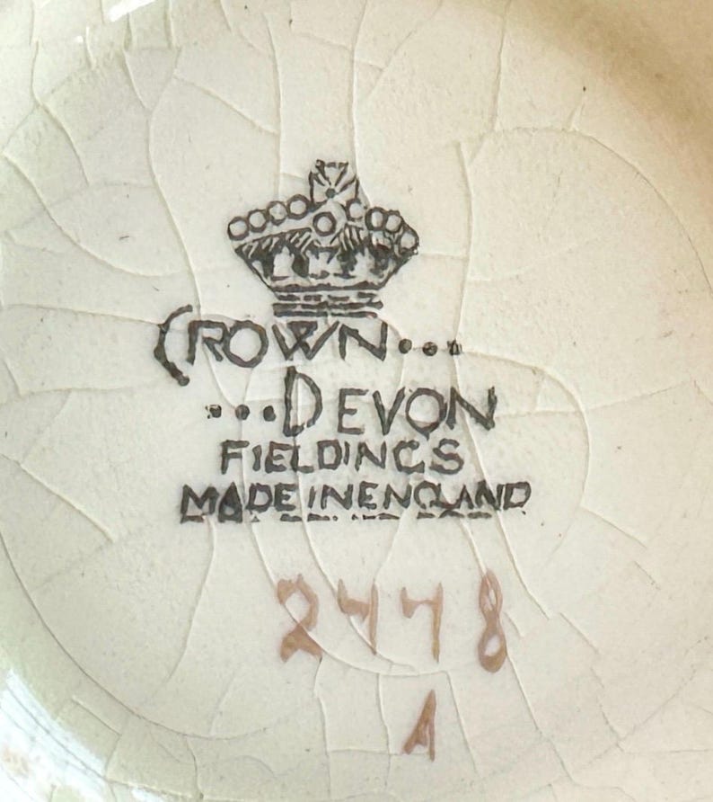 K&ouml;nnte beinhalten: Nahaufnahme einer cremefarbenen Keramikoberfl&auml;che mit Craquel&eacute;-Glasur. Das Bild zeigt den Text "CROWN... DEVON FIELDINGS MADE IN ENGLAND", die Zahlen "2478" und den Buchstaben "A". Ein schwarzes Kronenlogo ist ebenfalls zu sehen.