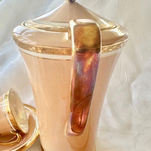 K&ouml;nnte beinhalten: Ein Vintage-Teeservice in Pfirsichfarbe mit goldenen Verzierungen. Das Set umfasst eine hohe Teekanne mit Deckel, einen Griff mit schillernden Akzenten und mehrere Tassen mit Untertassen. Das Design zeigt goldene B&auml;nder und einen klassischen, eleganten Stil.
