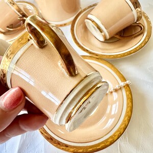 K&ouml;nnte beinhalten: Ein Set aus vintage Teetassen und Untertassen in Pfirsichfarbe mit goldenen Verzierungen. Die Tassen haben goldene Henkel und passende Untertassen. Eine Perlenkette liegt auf einer Untertasse. Das Set ist auf einer wei&szlig;en Oberfl&auml;che arrangiert.