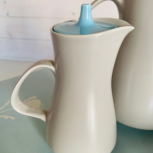May include: A cream-coloured ceramic creamer with a light blue lid. The creamer has a curved handle and a spout for pouring. A larger matching jug is partially visible in the background.