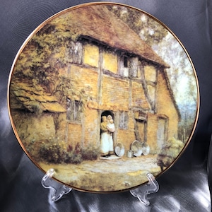 Puede incluir: Plato decorativo con una pintura detallada de una escena de cabaña con figuras en la puerta. El plato tiene un borde dorado y se exhibe en un soporte transparente. La obra de arte representa un edificio rústico con techo de paja.