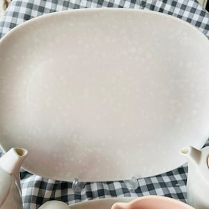 Op de afbeelding: Een grote, ovale, gebroken witte serveerschaal met een gespikkeld patroon. De schaal maakt deel uit van een theeservies, met theepotten en een melkkan zichtbaar. De achtergrond is een zwart-wit geblokt patroon.