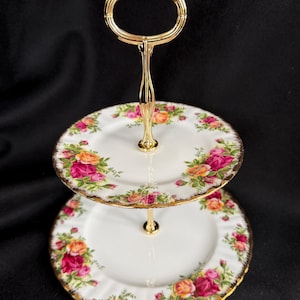 Puede incluir: Un soporte de dos niveles con platos de porcelana blanca de bordes festoneados, decorados con un patrón de rosas rosas y naranjas con hojas verdes. El soporte incluye un asa central y una varilla de conexión de metal dorado.