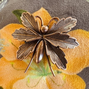 Peut inclure: Broche en métal de couleur bronze en forme d'insecte stylisé. La broche présente des ailes détaillées en forme de feuilles et un corps central avec des antennes. La broche est posée sur un fond à motifs floraux.