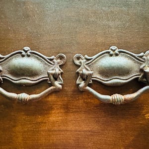 May include: A pair of antique drawer pulls with a distressed finish. Each pull has an ornate backplate and a curved handle. The metal has a weathered, tarnished appearance, suggesting age and use. The pulls are likely made of metal.