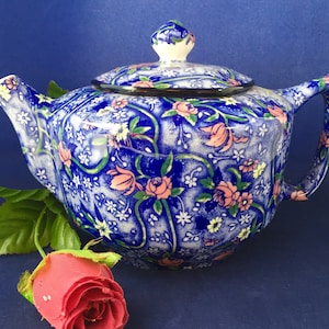 Könnte beinhalten: Eine blau-weiß gemusterte Teekanne mit passendem Deckel. Die Teekanne hat rosa und gelbe Blumen und grüne Ranken. Eine rote Rose und grüne Blätter sind im Vordergrund. Die Teekanne hat einen Henkel und einen Ausguss.