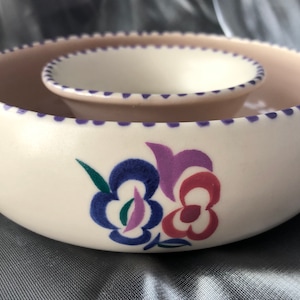 May include: Cream ceramic chip and dip serving set. The set includes a large bowl and a smaller bowl nested inside. The rims are decorated with purple dots. Floral designs in blue, purple, and red adorn the exterior.