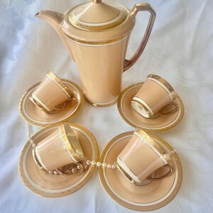 K&ouml;nnte beinhalten: Ein Vintage-Teeservice in Pfirsichfarbe mit goldenen Verzierungen. Das Set umfasst eine hohe Teekanne und vier Teetassen mit passenden Untertassen. Die Tassen und Untertassen sind um die Teekanne angeordnet. Eine Perlenkette ist &uuml;ber die Untertassen drapiert.