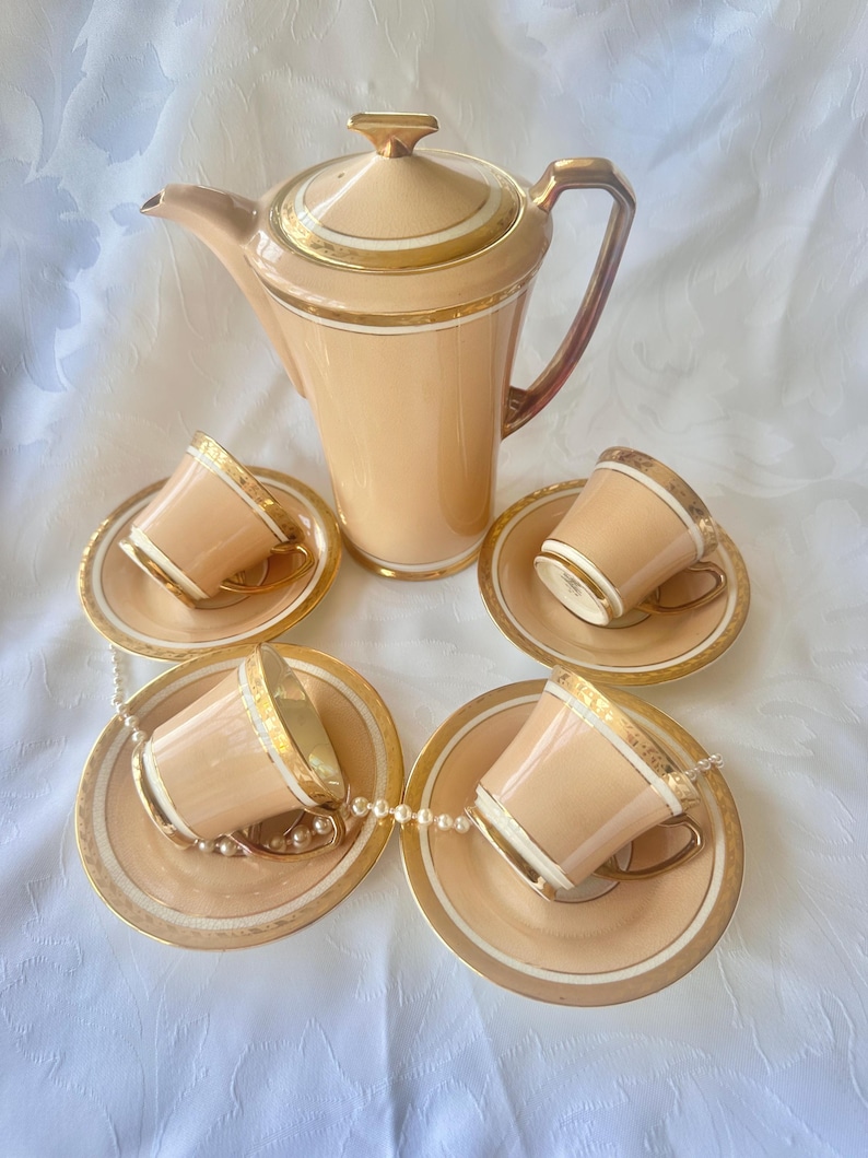 K&ouml;nnte beinhalten: Ein Vintage-Teeservice in Pfirsichfarbe mit goldenen Verzierungen. Das Set umfasst eine hohe Teekanne und vier Teetassen mit passenden Untertassen. Die Tassen und Untertassen sind um die Teekanne angeordnet. Eine Perlenkette ist &uuml;ber die Untertassen drapiert.