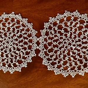 May include: Two white, circular doilies with intricate lace patterns. The doilies are placed side-by-side on a dark wooden surface, showcasing their delicate, floral-like designs. The lace appears to be hand-made, adding to their unique charm.