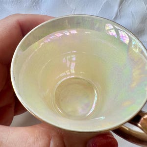 K&ouml;nnte beinhalten: Eine Vintage-Teetasse mit perlmuttfarbenem Innenraum und Goldrand. Das Innere der Tasse reflektiert einen Regenbogen von Farben. Der Griff hat eine warme, metallische Goldfarbe. Die Tasse wird von einer Person gehalten.