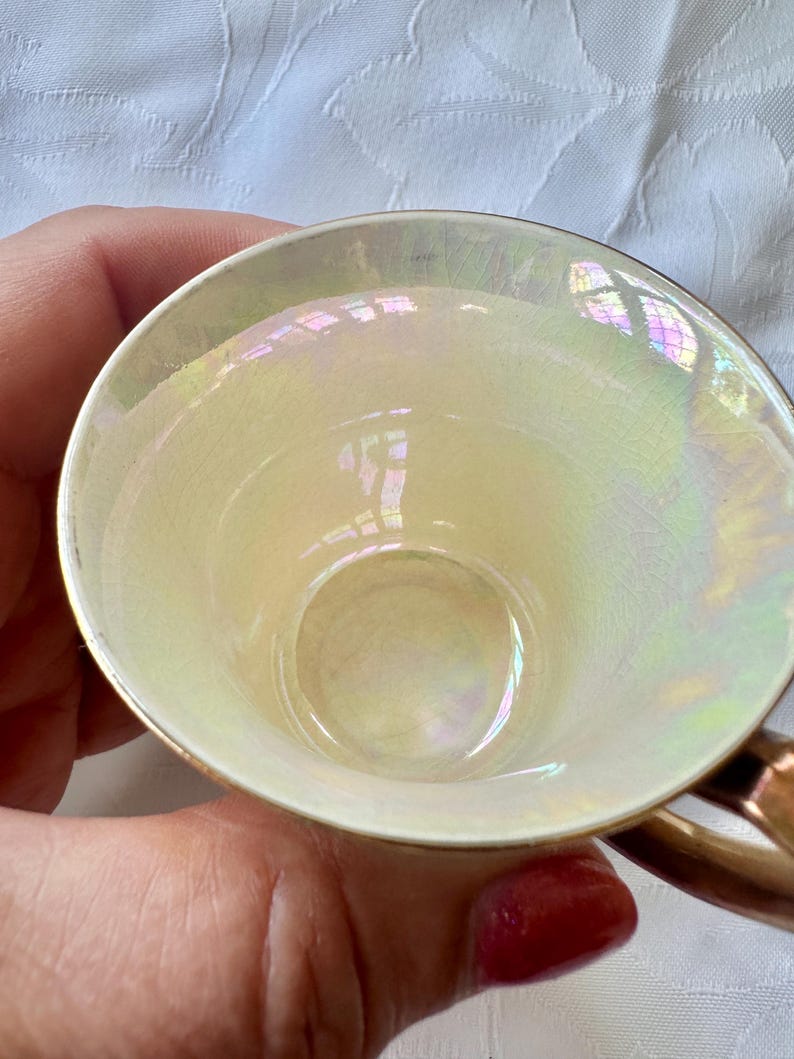 K&ouml;nnte beinhalten: Eine Vintage-Teetasse mit perlmuttfarbenem Innenraum und Goldrand. Das Innere der Tasse reflektiert einen Regenbogen von Farben. Der Griff hat eine warme, metallische Goldfarbe. Die Tasse wird von einer Person gehalten.