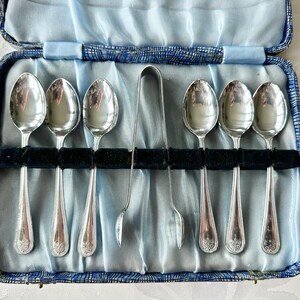 May include: A vintage set of six silver spoons and a sugar tong, displayed in a blue satin-lined case. The spoons feature a decorative handle design. The case is open, showing the contents.