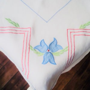 A pretty square vintage embroidered tablecloth, a blue flower, green leaves and red geometric accent in each corner on crisp white linen.