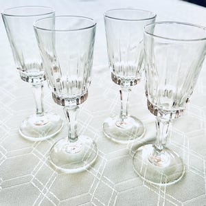 May include: Four clear stemmed glasses with a textured design on the bowl and a decorative band near the base. The glasses are arranged on a white tablecloth with a geometric pattern. The glasses are ideal for serving beverages.