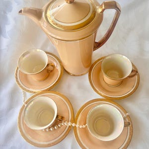 K&ouml;nnte beinhalten: Ein vintage-Teeservice in Pfirsichfarbe mit goldenen Verzierungen, bestehend aus einer Teekanne und vier Tassen mit Untertassen. Eine Perlenkette liegt &uuml;ber den Untertassen. Die Teekanne hat einen goldenen Griff und Deckel. Auf wei&szlig;em Untergrund.