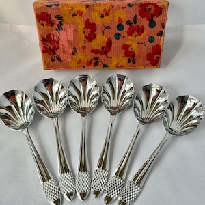 May include: A set of six silver-toned spoons with shell-shaped bowls and textured, cross-hatched handles, arranged in a row in front of a rectangular cardboard box featuring a vibrant floral pattern of red and yellow flowers on a peach background.