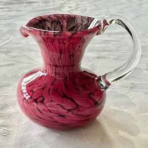 Peut inclure: Une cruche en verre soufflé à la main avec une poignée transparente. La cruche est d'un rouge vif avec des marbrures noires sur tout le corps. Le bord est évasé et la poignée est incurvée. Une pièce décorative pour la maison.