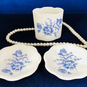 May include: A white ceramic tumbler and two small, scalloped dishes. Each piece is decorated with a blue floral pattern. A strand of white pearls is draped across the items against a dark blue background. The tumbler has a wavy top edge.