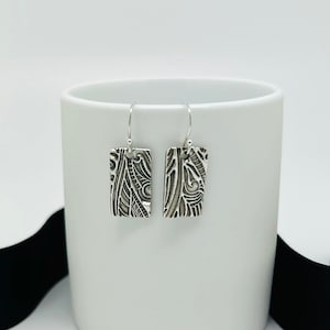 Peut inclure: Une paire de boucles d'oreilles en argent avec un motif texturé abstrait. Les boucles d'oreilles sont de forme rectangulaire et ont un style pendentif.