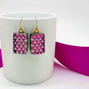 May include: A pair of rectangular earrings with a gold-colored hook. The earrings have a pink and purple textured design. The earrings are displayed on a white mug with a fuchsia ribbon in the background.