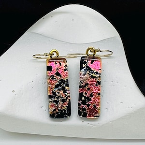 May include: A pair of rectangular dangle earrings with a gold-tone hook. The earrings feature a black base with pink and gold accents. The earrings are displayed on a white surface with a black ribbon in the background.
