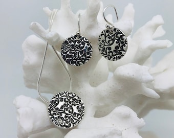 May include: A silver jewelry set featuring a pendant necklace and matching earrings. Each piece has a circular design with a black, swirling pattern. The jewelry is displayed on a white, coral-like structure, creating a striking contrast.