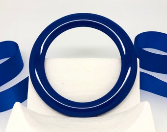 May include: A glossy, navy blue bangle bracelet. The circular bracelet is displayed on a white surface with a blue ribbon in the background. The bangle is a solid color and has a smooth, reflective surface.