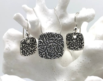 May include: A silver jewelry set featuring a square pendant necklace and matching earrings. The pendant and earrings have a textured, organic pattern. The set is displayed on a white coral-like structure, creating a striking contrast.