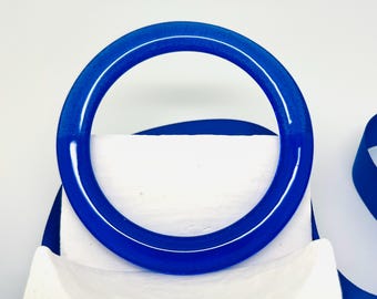 May include: A vibrant blue bangle bracelet. The circular bracelet is a solid, glossy blue with a white highlight. The bracelet is displayed on a white surface with a blue ribbon in the background. The bangle is a simple, elegant accessory.