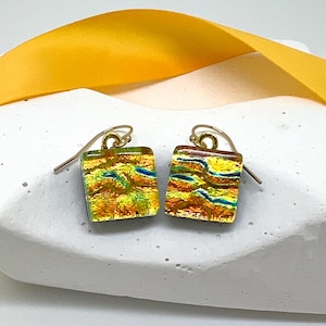 May include: A pair of square dichroic glass earrings with gold-colored ear wires. The earrings feature a blend of yellow, orange, and blue colors, creating a textured, layered effect. A yellow ribbon is in the background.