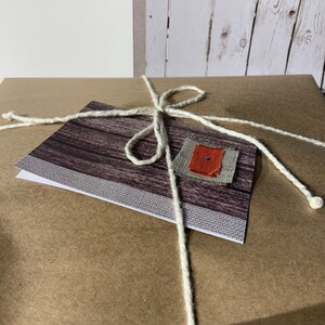 May include: A square package wrapped in brown paper and tied with a white string. A rectangular card with a dark wood grain pattern and a small square fabric patch in gray and orange is secured to the top of the package by the white string.